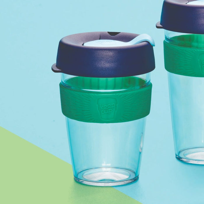 KeepCup Original Clear M 12oz in Eventide, featuring a transparent aqua-tinted cup with green silicone band and navy blue lid, shown on a light blue and lime green background with a second cup partially visible.