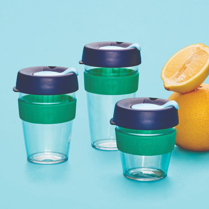 Three KeepCup Original Clear reusable cups in the Eventide colourway, shown in varying sizes with clear plastic bodies, green silicone bands and navy lids, arranged on a light blue background alongside fresh oranges and a halved lemon.