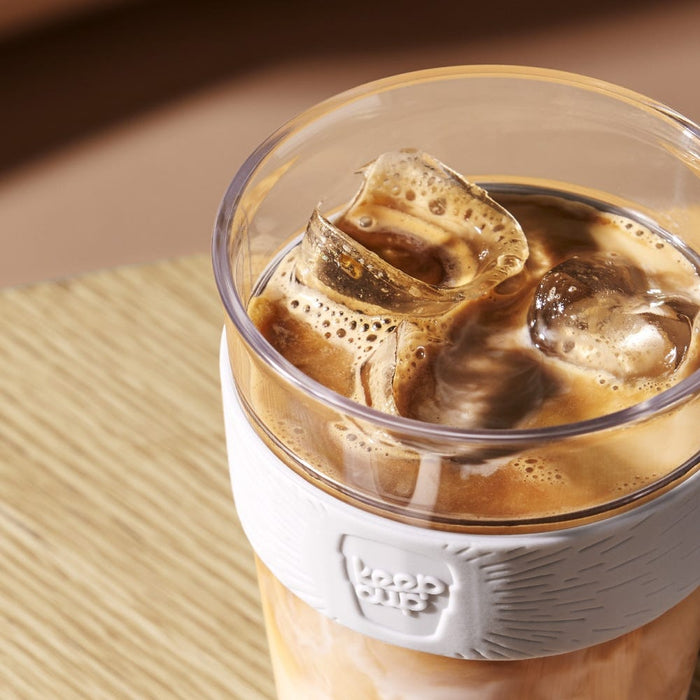 KeepCup Original Clear S 8oz in Moonshine, featuring a plastic cup with white silicone band, filled with iced coffee and visible ice cubes, photographed from above on a natural woven placemat.