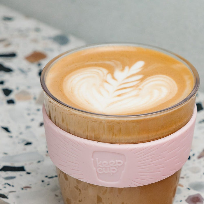A KeepCup Original Clear M 12oz in Peaches sits on a terrazzo surface, featuring a plastic cup with a soft pink silicone band and filled with a latte displaying a heart-shaped rosetta pattern in the foam.