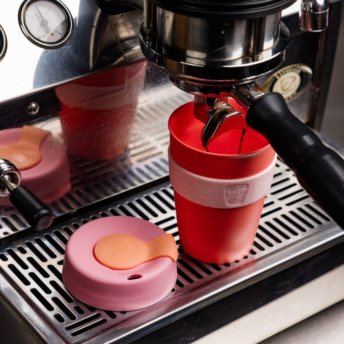 A KeepCup Original M 12oz in Peaches sits on an espresso machine drip tray with espresso pouring into it from a portafilter, its pink and peach lid placed beside it.
