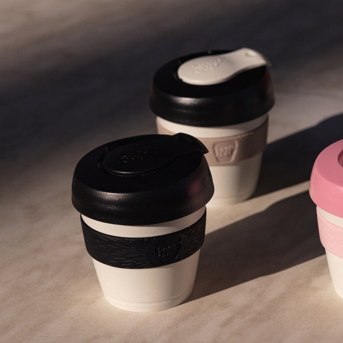 KeepCup Original XXS 4oz in Raven, featuring a white cup with black lid and black band, displayed on a concrete surface alongside two other KeepCup colour variations in natural lighting.