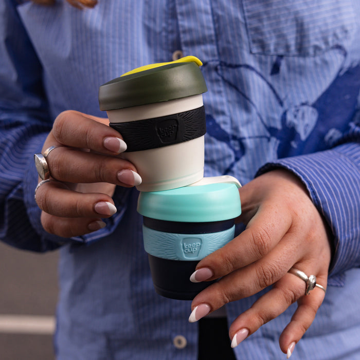 A person in a blue striped shirt holds two stacked KeepCup reusable coffee cups, with the Original XXS 4oz Pine cup on top featuring its distinctive olive green lid with yellow plug, cream body and black band, balanced above a dark navy and mint coloured cup.