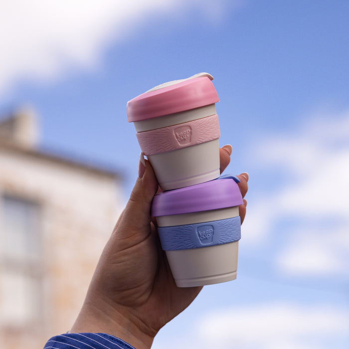 Hand holding two stacked KeepCup Original XXS 4oz reusable cups against a blue sky, featuring the Lilac colourway with purple lid and blue band on the bottom cup, and a pink-lidded cup on top.