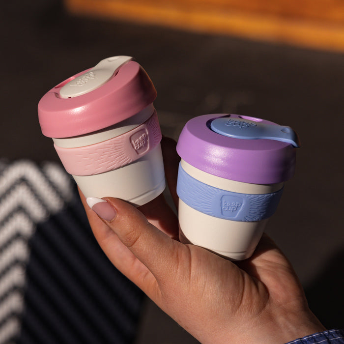 A hand with white nail polish holds two KeepCup Original reusable cups – the XXS 4oz Lilac colourway on the right featuring a purple lid, lavender band and cream body, alongside a pink and white version on the left.