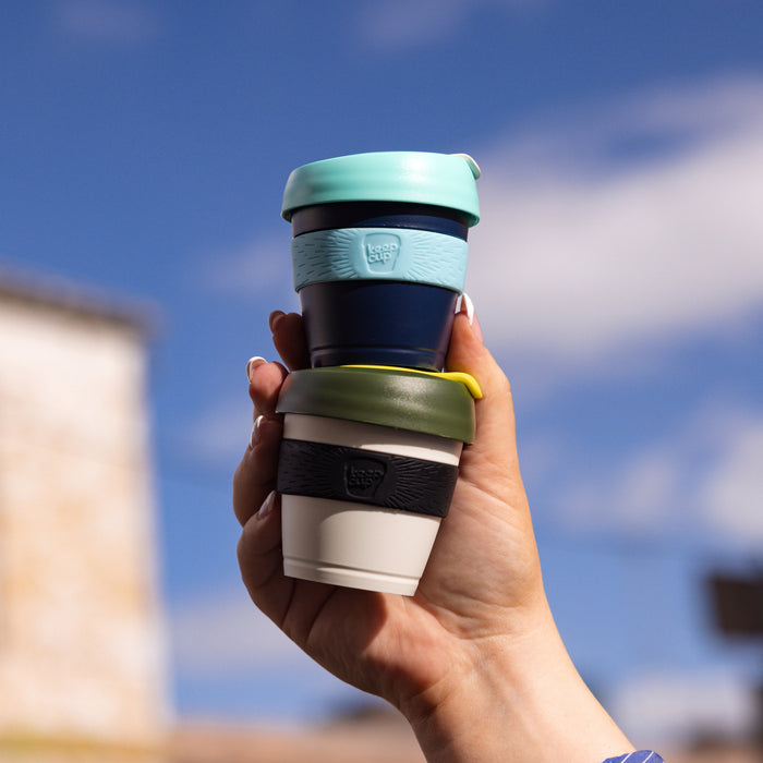 A hand holds two stacked KeepCup reusable coffee cups against a bright blue sky, featuring the Original XXS 4oz Pine cup in olive green with a yellow band on the bottom, and a navy cup with mint green lid and band on top.