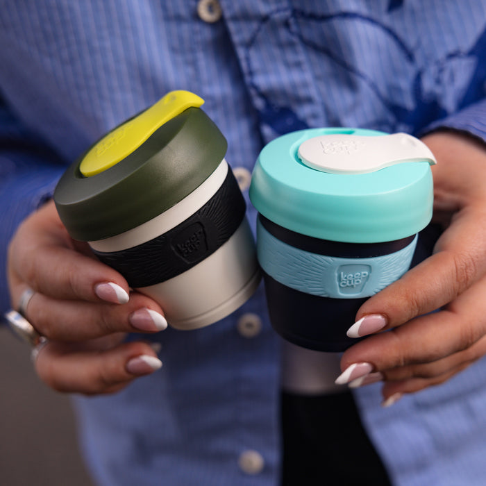 Two hands with white-tipped nails holding KeepCup reusable coffee cups, featuring the Original XXS 4oz Pine on the left with its olive green lid, yellow plug and black band, alongside a turquoise and navy cup on the right, against a blue shirt background.