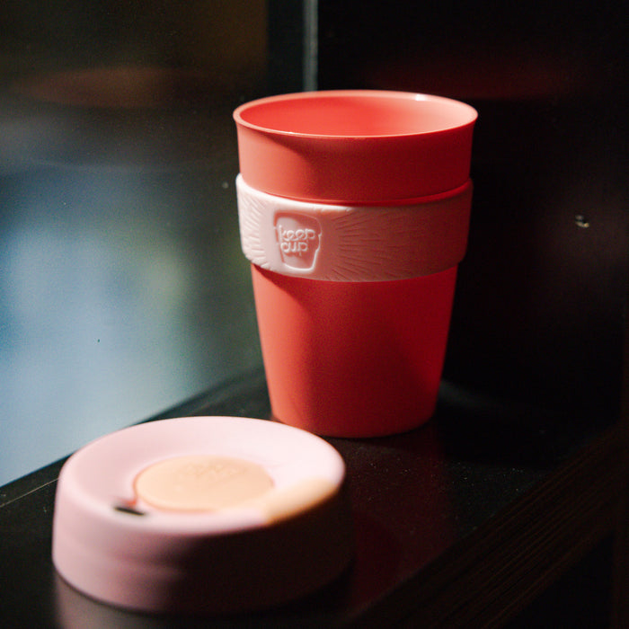 KeepCup Original in coral with a pale pink lid and band, displayed on a dark shelf with the lid resting beside the cup, showcasing the silicone grip band with embossed KeepCup logo.