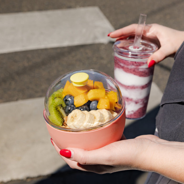 A person with red nail polish holds the KeepCup Go Bowl Original in Clay, filled with fresh fruit including kiwi, blueberries, mango and banana slices, while their other hand holds a layered berry smoothie in a clear tumbler, seated outdoors on concrete steps.