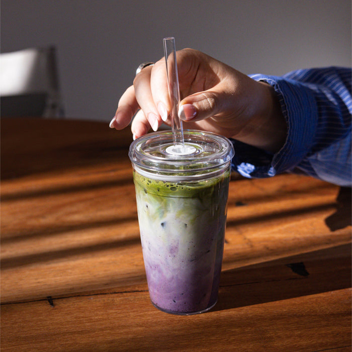 A hand with manicured nails holds a clear straw inserted into the Urban Adventurer Black KeepCup, which sits on a wooden table and contains a layered purple and green iced drink, likely a taro and matcha latte.
