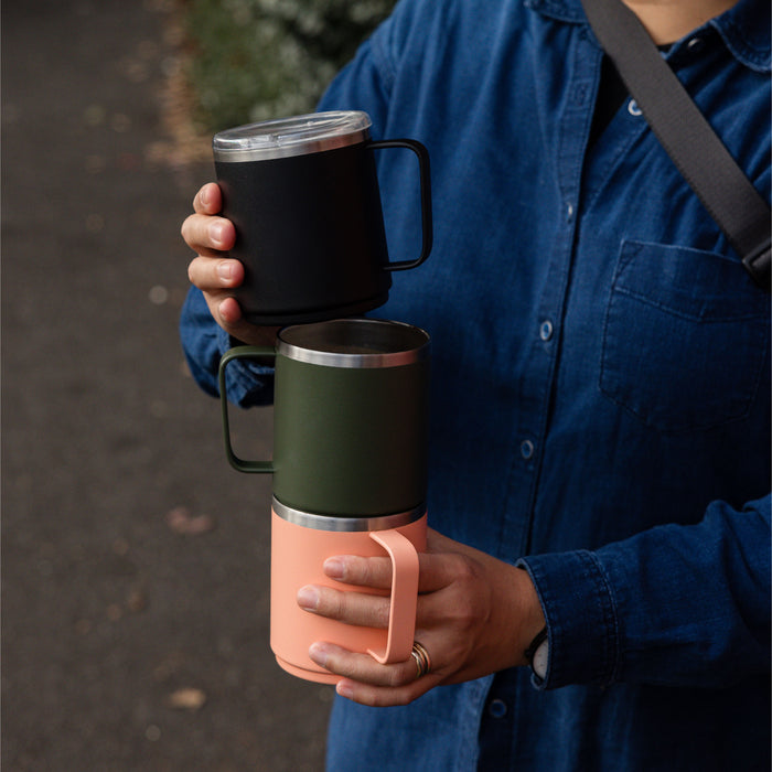 A person in a blue denim shirt holding three stacked KeepCup Urban Adventurer insulated mugs in Black, olive green, and coral, showcasing the stackable design with matching coloured handles and stainless steel rims.