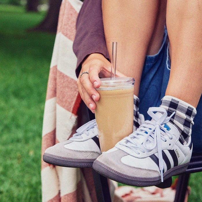 A hand holds the Urban Adventurer Black KeepCup filled with iced coffee, featuring a clear lid and reusable straw, with a casual outdoor park setting and Adidas Samba sneakers visible in the background.