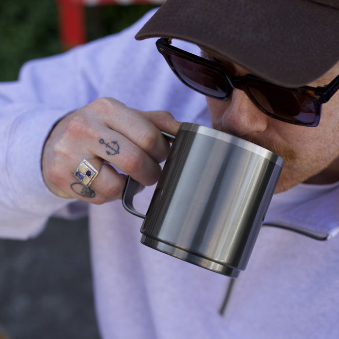 A man wearing sunglasses and a cap takes a sip from a KeepCup Camp Mug M 12oz in Nitro, showcasing the brushed stainless steel finish and sturdy handle, with a small anchor tattoo visible on his finger.