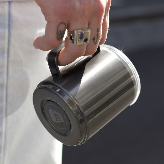 A tattooed hand with a silver ring holds the KeepCup Camp Mug M 12oz in Nitro by its black handle, showing the brushed stainless steel body and dark base with KeepCup branding.