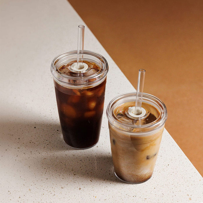 Two KeepCup Urban Adventurer Nitro reusable cups in different sizes on a speckled stone surface, the larger filled with black iced coffee and the smaller with iced latte, both featuring clear double-walled construction, lids and plastic straws.
