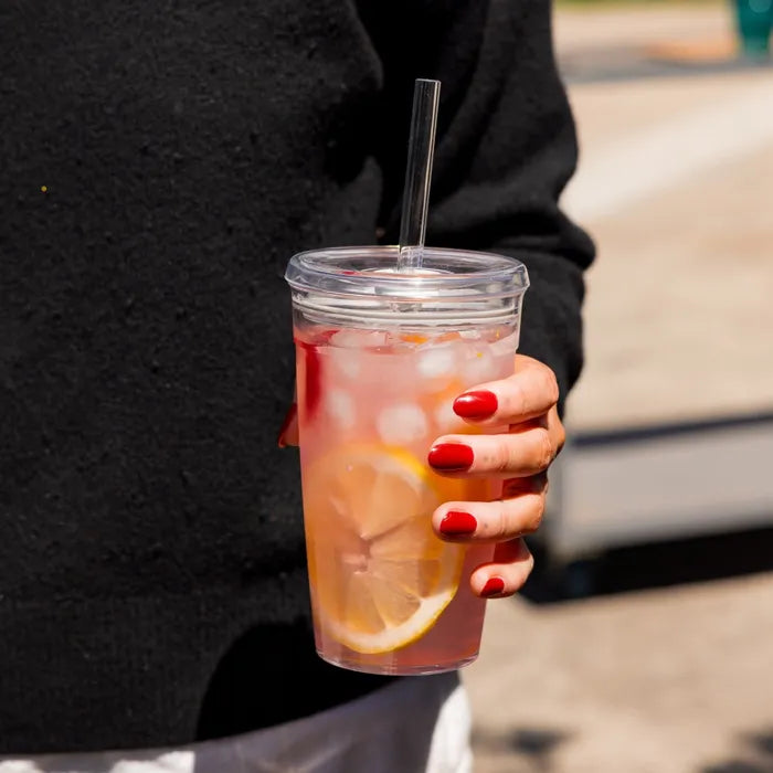 Hand with red nail polish holding a Cold Cup Original L 16oz filled with iced tea, lemon slices and ice, with a Coffee Straw, against a black jumper in an outdoor setting.