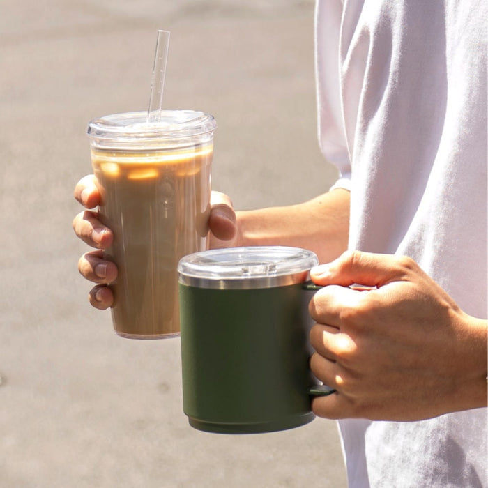 A person holding two KeepCup products outdoors — a clear double-walled cup with iced coffee and straw in one hand, and the Urban Adventurer in Pine, a dark green insulated mug with clear lid, in the other.